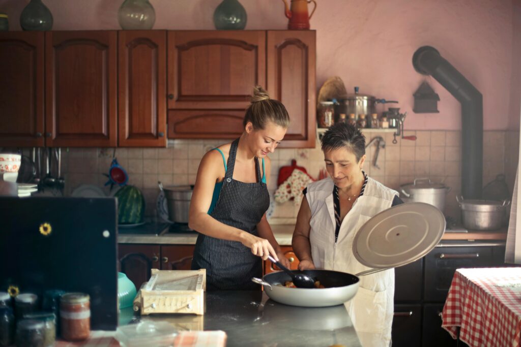 pexels-photo-3772534-3772534 Daughter and senior mother standing at table in kitchen and stirring dish in frying pan while preparing food for dinner
